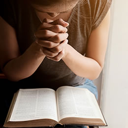 A woman seated with hands clasped together reading the bible
