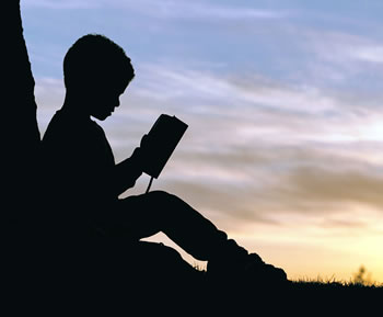 Silhouette of a boy sitting against a tree reading a book.