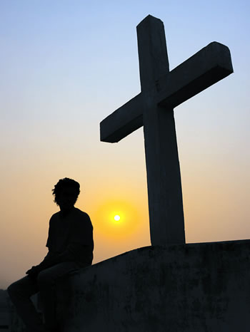 Silhoutte of a person sitting on a hill by a cross.