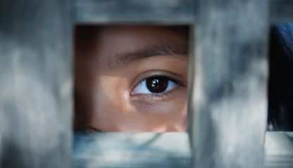 The blank stare of a child's eye who is standing behind wooden cage.